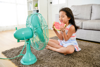 Small female child sitting in front of electric fan blowing cooling wind and eating cold watermelon feeling refreshing during summer season.