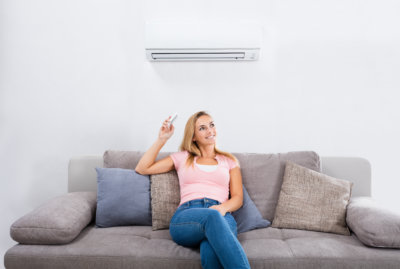 Young Happy Woman Sitting On Couch Operating Air Conditioner With Remote Control At Home