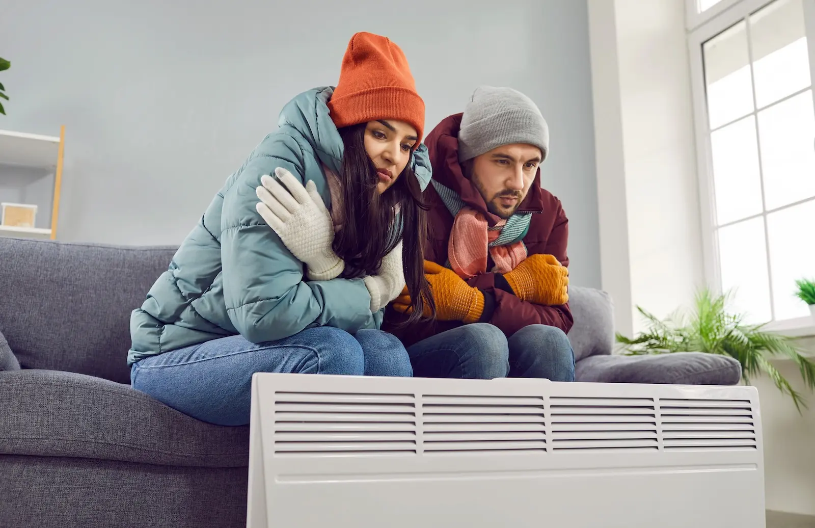 Freezing couple sitting on couch with layers and space heater.