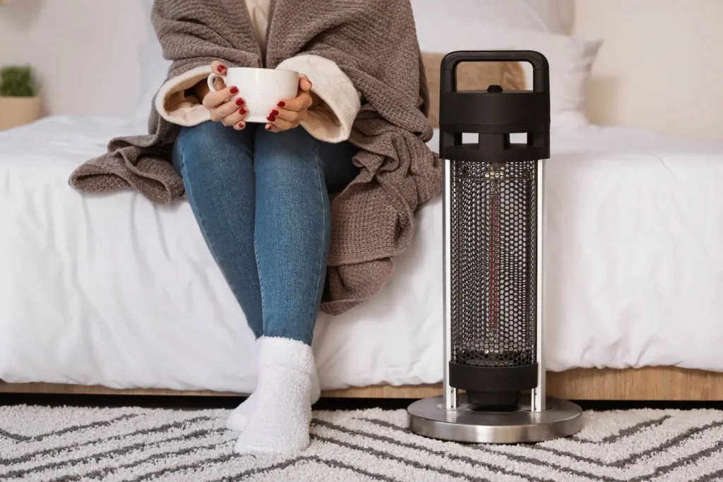 A person wrapped in a blanket sitting on a bed, holding a mug next to a small space heater on a rug in a cozy bedroom.